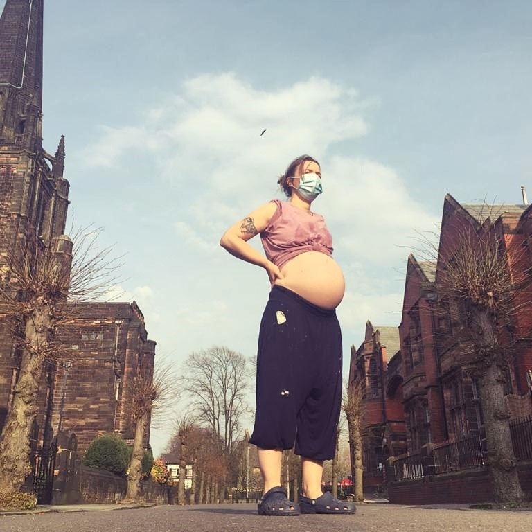 LHL_2020_001: Photograph depicting Becky Crawford who stands outside St George’s church, Davenport. They wear a surgical mask, a pink sleeveless top, a pair of navy cropped trousers and a pair of Crocs. The top is pulled up to reveal the bump.