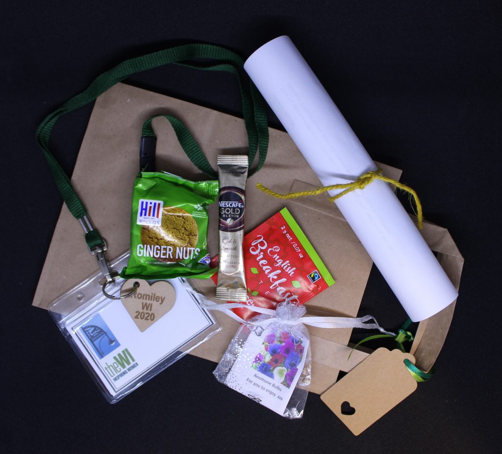 LHL_2020_007b: a photograph of a number of items lay on a brown paper bag. They are a packet of ginger nut biscuits, a sachet of coffee, an English Breakfast tea bag, a rolled up piece of paper tied with yellow string, a mesh bag containing a plant bulb, and a green lanyard attached to a plastic name tag.