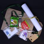 LHL_2020_007b: a photograph of a number of items lay on a brown paper bag. They are a packet of ginger nut biscuits, a sachet of coffee, an English Breakfast tea bag, a rolled up piece of paper tied with yellow string, a mesh bag containing a plant bulb, and a green lanyard attached to a plastic name tag. Thumbnail