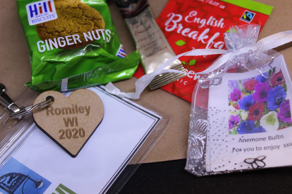 LHL_2020_007c: a close up photograph of the items lay on a brown paper bag - a packet of ginger nut biscuits, a sachet of coffee, an English Breakfast tea bag, a mesh bag containing a plant bulb, and the name tag which has a wooden heart-shaped keyring charm, stamped with 'Romiley WI 2020'.