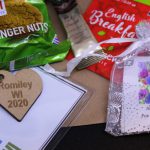 LHL_2020_007c: a close up photograph of the items lay on a brown paper bag - a packet of ginger nut biscuits, a sachet of coffee, an English Breakfast tea bag, a mesh bag containing a plant bulb, and the name tag which has a wooden heart-shaped keyring charm, stamped with 'Romiley WI 2020'. Thumbnail