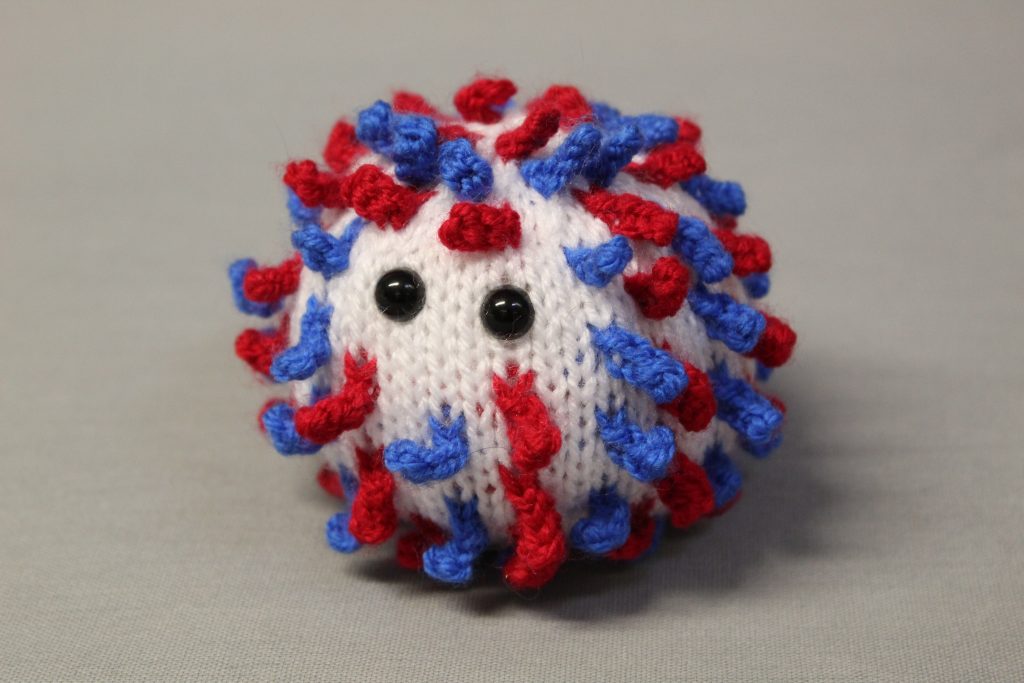 STOPM_2020_452a: Image of knitted toy or souvenir of an anthropomorphic coronavirus; white spherical shape with blue and red 'spikes' to mimic the structure of the coronavirus particle. Two black plastic domed beads/buttons have been added as 'eyes'. The coronavirus was knitted in red, white and blue to commemorate the 75th anniversary of VE (Victory in Europe) Day which was celebrated on 8th May 2020. Knitted by Linda Cornes Parker, who is a part of the group Knitting Friends in the Park.; knitted toy or souvenir of an anthropomorphic coronavirus; white with multicoloured speckles spherical shape with 'spikes' to mimic the structure of the coronavirus particle. Two black plastic domed beads/buttons have been added as 'eyes'.  A wooden cocktail stick and a wooden parallelogram have been cello taped and stuck into the ball as a sign which has been written on in black pen 'WASH YOUR HANDS'. Knitted by Linda Cornes Parker, who is a part of the group Knitting Friends in the Park.