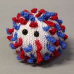 STOPM_2020_452a: Image of knitted toy or souvenir of an anthropomorphic coronavirus; white spherical shape with blue and red 'spikes' to mimic the structure of the coronavirus particle. Two black plastic domed beads/buttons have been added as 'eyes'. The coronavirus was knitted in red, white and blue to commemorate the 75th anniversary of VE (Victory in Europe) Day which was celebrated on 8th May 2020. Knitted by Linda Cornes Parker, who is a part of the group Knitting Friends in the Park.; knitted toy or souvenir of an anthropomorphic coronavirus; white with multicoloured speckles spherical shape with 'spikes' to mimic the structure of the coronavirus particle. Two black plastic domed beads/buttons have been added as 'eyes'.  A wooden cocktail stick and a wooden parallelogram have been cello taped and stuck into the ball as a sign which has been written on in black pen 'WASH YOUR HANDS'. Knitted by Linda Cornes Parker, who is a part of the group Knitting Friends in the Park. Thumbnail