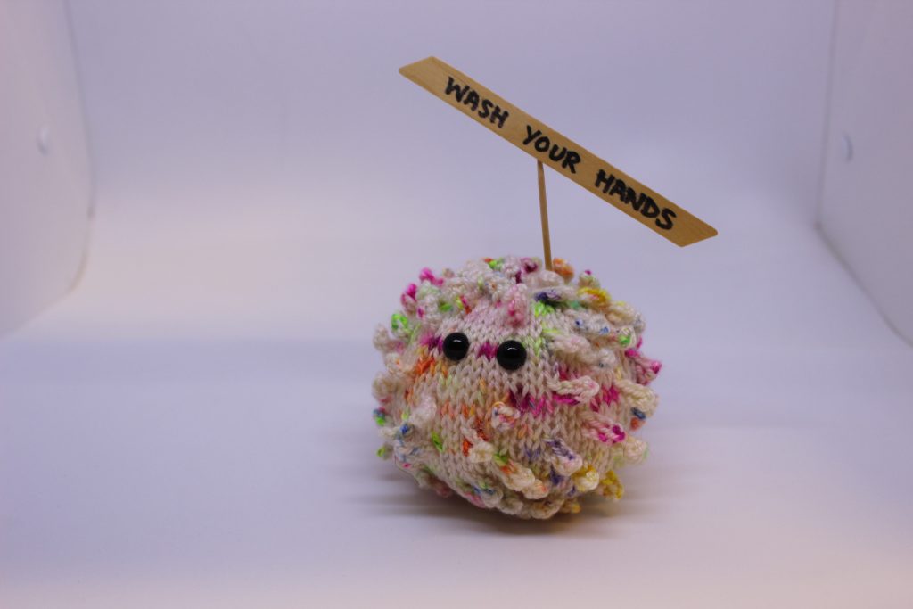 STOPM_2020_453a: knitted toy or souvenir of an anthropomorphic coronavirus; white with multicoloured speckles spherical shape with 'spikes' to mimic the structure of the coronavirus particle. Two black plastic domed beads/buttons have been added as 'eyes'.  A wooden cocktail stick and a wooden parallelogram have been cello taped and stuck into the ball as a sign which has been written on in black pen 'WASH YOUR HANDS'. Knitted by Linda Cornes Parker, who is a part of the group Knitting Friends in the Park. 