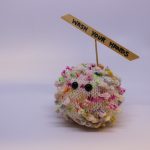 STOPM_2020_453a: knitted toy or souvenir of an anthropomorphic coronavirus; white with multicoloured speckles spherical shape with 'spikes' to mimic the structure of the coronavirus particle. Two black plastic domed beads/buttons have been added as 'eyes'.  A wooden cocktail stick and a wooden parallelogram have been cello taped and stuck into the ball as a sign which has been written on in black pen 'WASH YOUR HANDS'. Knitted by Linda Cornes Parker, who is a part of the group Knitting Friends in the Park.  Thumbnail