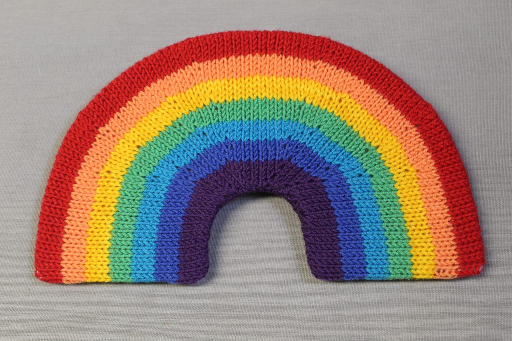 STOPM_2020_454a: knitted toy or souvenir of a rainbow; arch-shaped piece of flexible plastic, covered with a knitted rainbow in the colours of red, orange, yellow, green, turquoise, blue, and purple. Knitted by Linda Cornes Parker, who is a part of the group Knitting Friends in the Park.