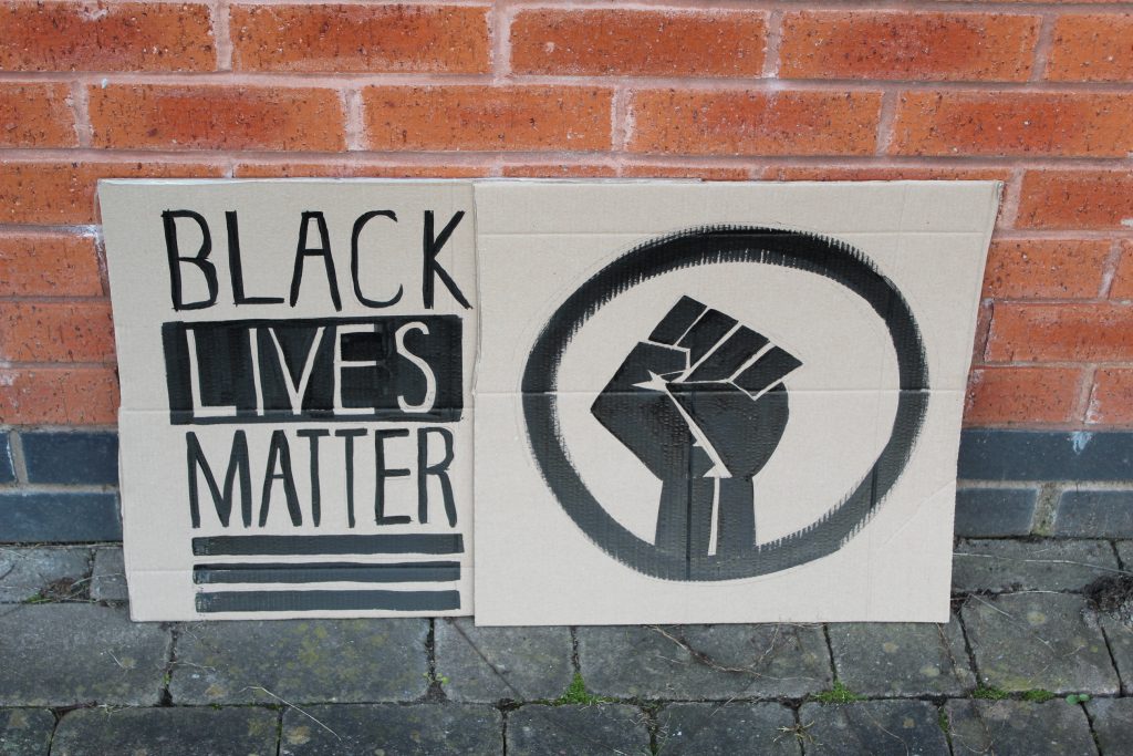 STOPM_2020_455_456a: Photo of two home made placards, stood in front of a red brick building.  The signs have been made from sections of a cardboard box. One sign has been drawn and painted with the words 'BLACK LIVES MATTER', and the other has been painted with the symbol of a black raised fist in a circle. The clenched fist has become synonymous with the Black Lives Matter movement but has a long history.