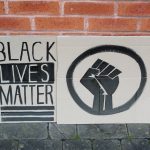 STOPM_2020_455_456a: Photo of two home made placards, stood in front of a red brick building.  The signs have been made from sections of a cardboard box. One sign has been drawn and painted with the words 'BLACK LIVES MATTER', and the other has been painted with the symbol of a black raised fist in a circle. The clenched fist has become synonymous with the Black Lives Matter movement but has a long history. Thumbnail