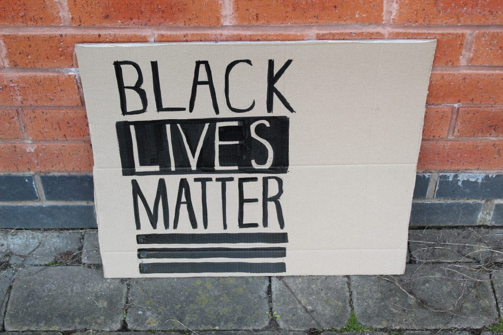 STOPM_2020_455a: Photo of a home made placard, stood in front of a red brick building. The sign has been made from a section of a cardboard box.  The sign has been drawn and painted with the words 'BLACK LIVES MATTER' in black paint. 
