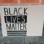 STOPM_2020_455a: Photo of a home made placard, stood in front of a red brick building. The sign has been made from a section of a cardboard box.  The sign has been drawn and painted with the words 'BLACK LIVES MATTER' in black paint.  Thumbnail
