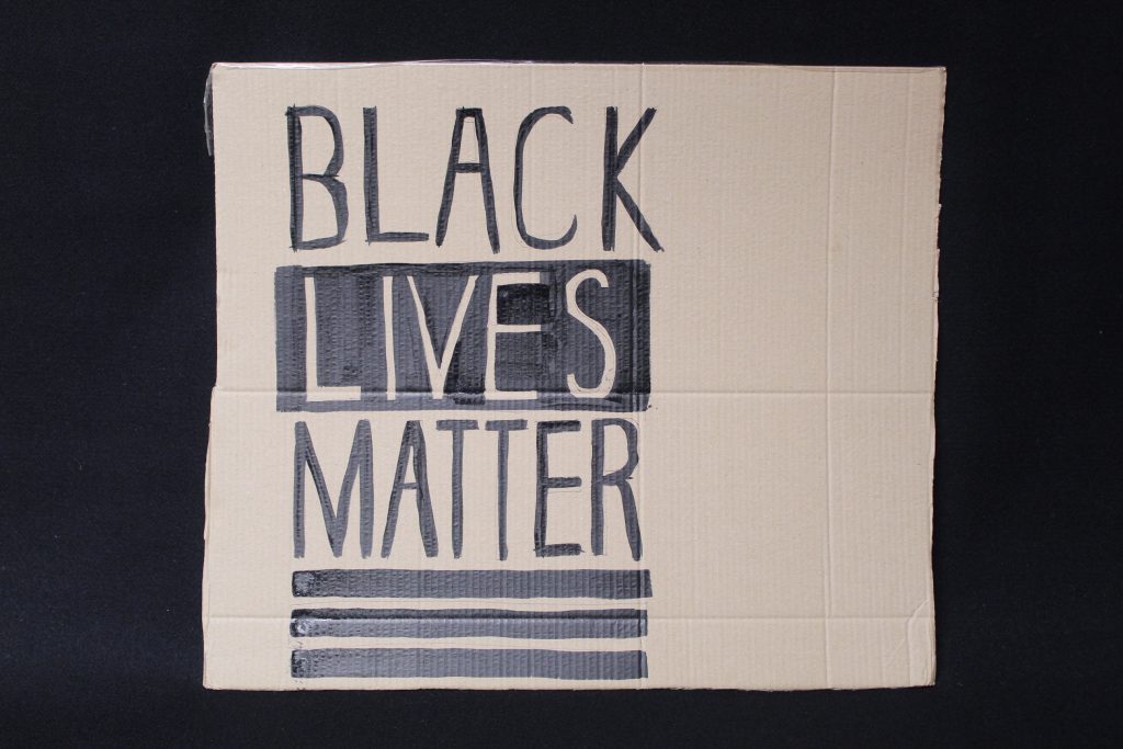 STOPM_2020_455b: Photo of a home made placard, on a black background. The sign has been made from a section of a cardboard box.  The sign has been drawn and painted with the words 'BLACK LIVES MATTER' in black paint