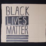 STOPM_2020_455b: Photo of a home made placard, on a black background. The sign has been made from a section of a cardboard box.  The sign has been drawn and painted with the words 'BLACK LIVES MATTER' in black paint Thumbnail
