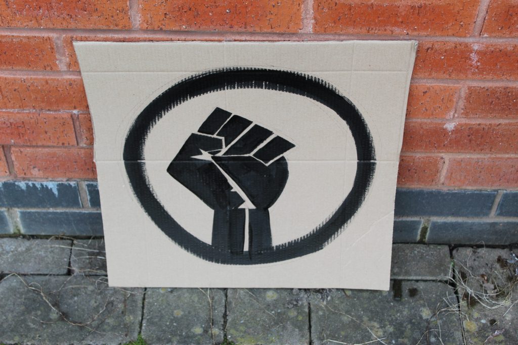 STOPM_2020_456a: Photo of a home made placard, stood in front of a red brick building. The sign has been made from a section of a cardboard box.  The sign has been painted with the symbol of a black raised fist in black paint.