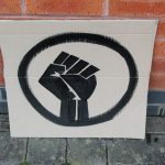 STOPM_2020_456a: Photo of a home made placard, stood in front of a red brick building. The sign has been made from a section of a cardboard box.  The sign has been painted with the symbol of a black raised fist in black paint. Thumbnail