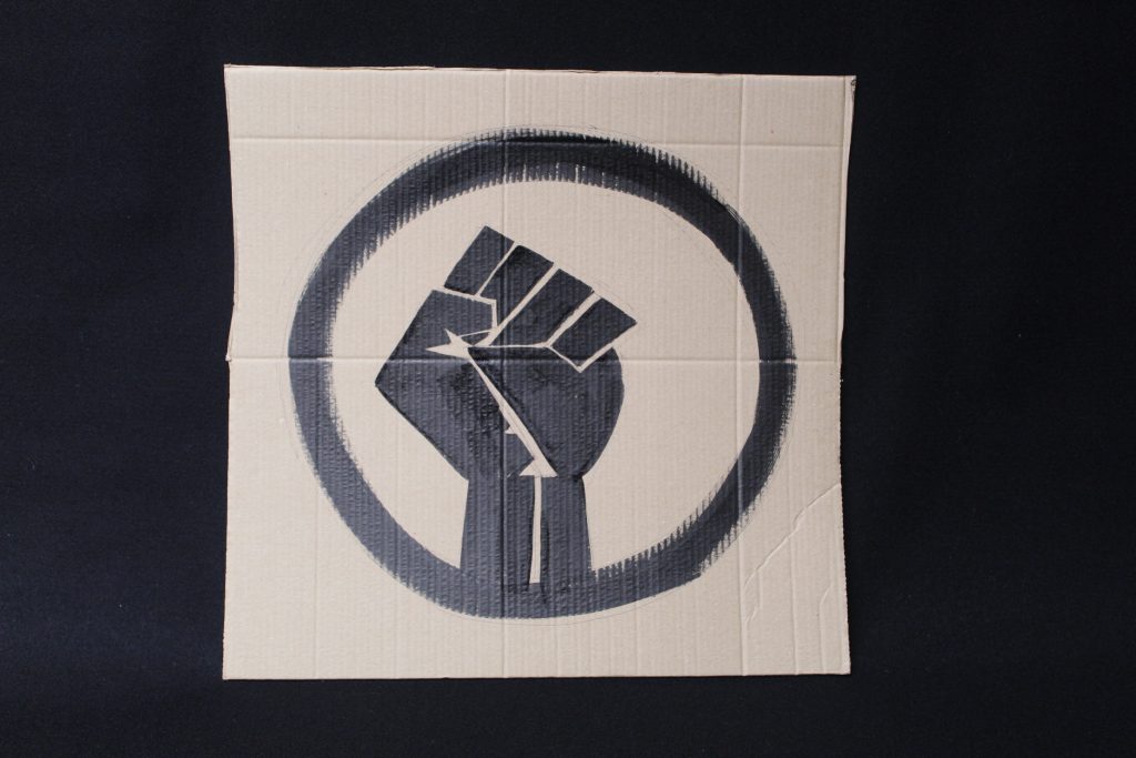 STOPM_2020_456b: Photo of a home made placard, on a black background. The sign has been made from a section of a cardboard box.  The sign has been painted with the symbol of a black raised fist in black paint.